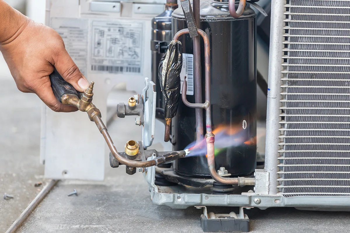 An HVAC technician uses a brazing torch with a blue flame to repair or solder copper lines on an outdoor air conditioning unit compressor, demonstrating a crucial step in regular HVAC maintenance to prevent system leaks