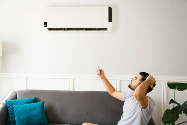 A man sits on a couch, raising his hand toward the AC unit above him, frustrated because the AC compressor is not turning on.