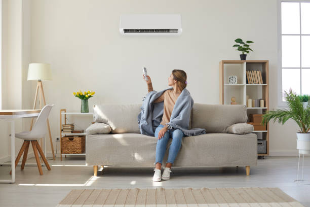 A woman sits on a couch, looking concerned in front of a white ac running but not cooling.