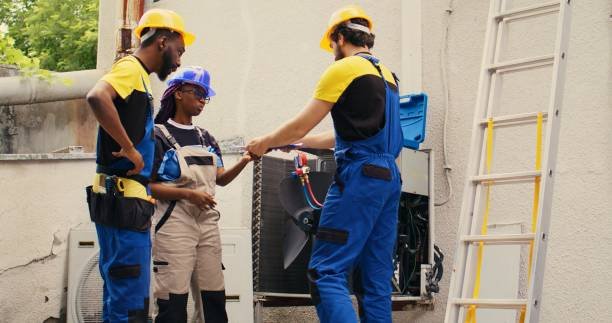 Three men in blue overalls and hard hats work together on a garage AC installation, mounting the air conditioning unit on a wall to ensure proper cooling.