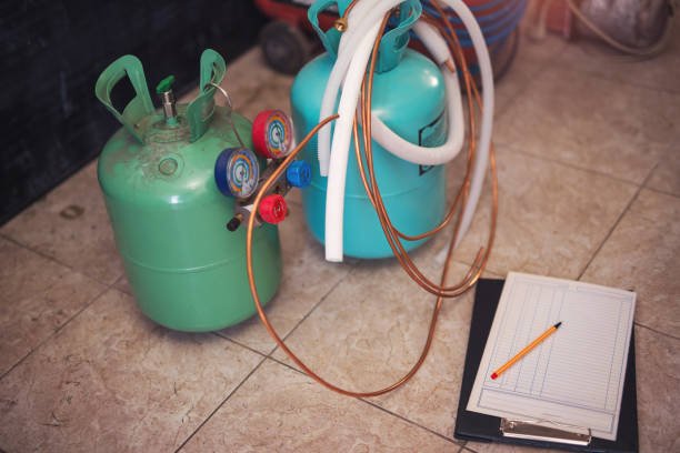 A gas cylinder, a hose, and a notebook are arranged on a tile floor, indicating a refrigerant recharge service setup.