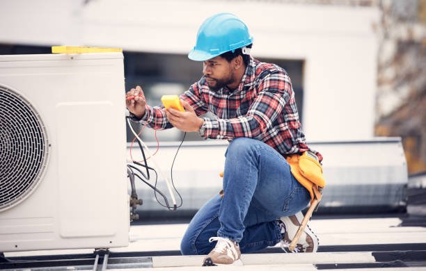 A man in a hard hat and blue shirt repairs an air conditioner as part of an AC servicing task.