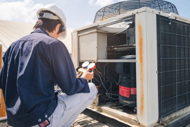 A man in a hard hat and white shirt repairs an air conditioner as part of a heating repair system.