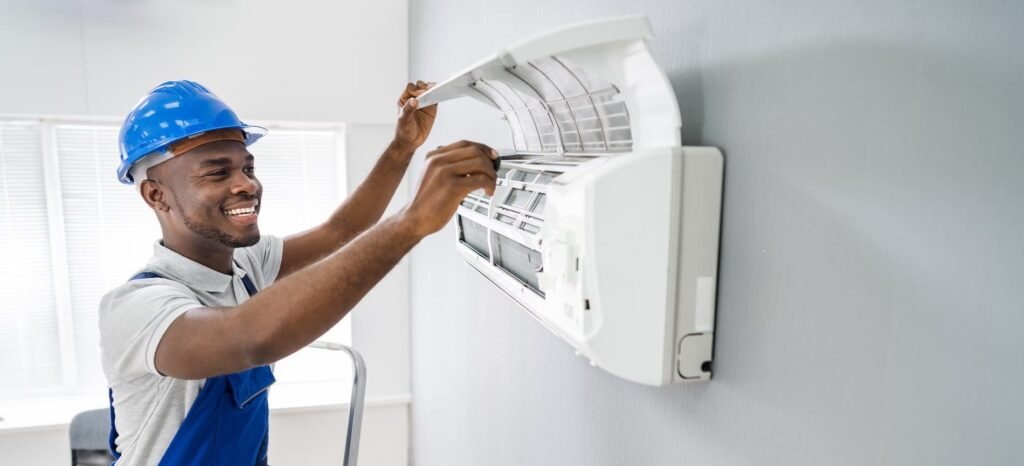 A man in a hard hat and blue shirt repairs a residential air conditioner on a home exterior.